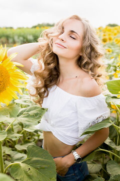 a young girl with a model appearance, blond and long hair, in denim trousers and a white blouse in a sunflower field