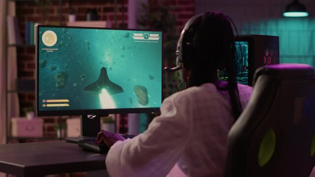 Over shoulder view of african american gamer girl playing space shooter simulation using pc gaming setup enjoying free time at home. Woman streaming online action game multiplayer competition.