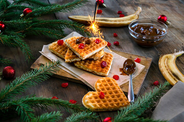 Homemade Belgian waffles heart shaped on plate with colorful red sweet cherry berries, banana, and boiled condensed milk,flaming bengal fire. Christmas new year background,dessert food - obrazy, fototapety, plakaty
