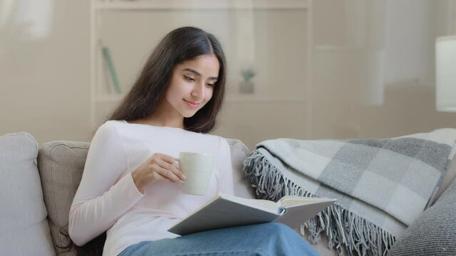 Serene young girl student daughter relaxed arabian biracial intelligent woman sits on sofa reads paper book scientific literature educational textbook preparing for homework exams enjoying bestseller