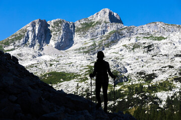 Fototapeta premium Silhouette of Woman Hiker Standing on the Edge of a Mountain Trail and Admiring The Krn Mountain