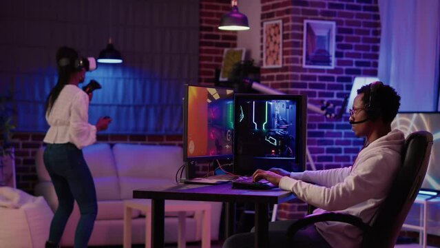 Gamer playing multiplayer first person shooter while gaming girl is fighting in virtual reality boxing game. Man streaming online action game on pc while girlfriend uses vr goggles for simulation.