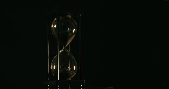Rotation of sand timer clock with sand flowing from through hourglass on black background.
