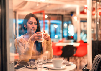 young latin woman sitting alone holding a cup of coffee or tea in a cafe seen through the window