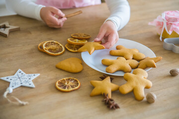 A child's hand takes from a white plate Christmas homemade cookies in the shape of a star, On a wooden table lie cookies gingerbread man, dried orange, anise, peenium molds and a white forfor cup