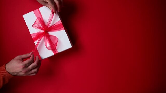 Person opening gift box with red ribbon. Top view of hands opening Christmas present. Red background.Merry Christmas.Winter holidays,New Year,birthday presents. Vertical 4K footage for reels, stories