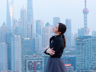 Beautiful young Chinese woman in black arms crossed on top of mansion roof with blur Shanghai Bund landmark buildings background with dusk light. Emotions, people, beauty and lifestyle concept.
