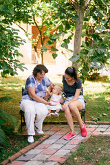 Mom, grandmother and little girl are sitting on a wooden bench in the park