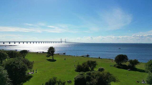 Oresundsbron, Oresund Bridge Over The Glistening Waters Of Oresund From The Park With People In Limhamn, Sweden. - aerial pullback