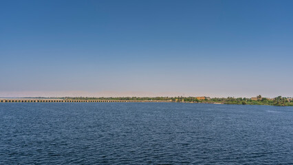 The dam of the sluice system in Esna. A long concrete bridge crosses a wide river. Green vegetation is visible far away on the shore. Clear blue sky. Copy space. Egypt. Nile