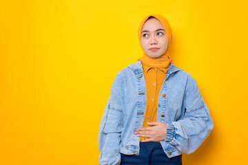 Portrait of young Asian woman in jeans jacket keep hands on the stomach and feel satiety after eating tasty isolated over yellow background