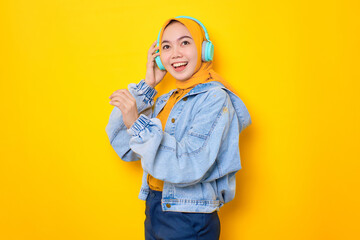 Dancing young Asian woman in jeans jacket wearing headphones for listening to music isolated over yellow background