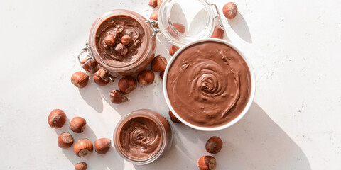 Jars and bowl with tasty chocolate paste and hazelnuts on light background, top view