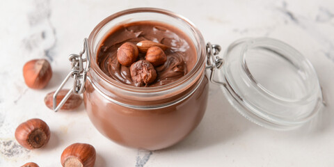 Jar with tasty chocolate paste and hazelnuts on light background, closeup