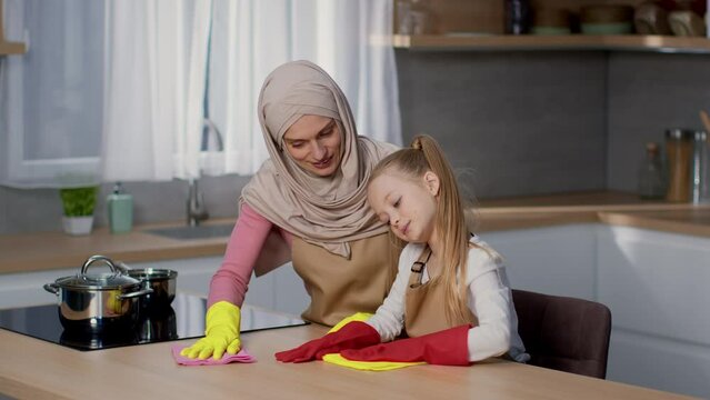 Domestic chores. Happy muslim mother in hijab and her cute little daughter cleaning table at kitchen, empty space