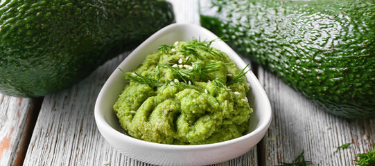 Bowl with tasty guacamole and ripe avocados on wooden table, closeup