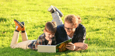 Father and his little son reading book while lying on green grass in park