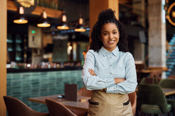 Happy African American waitress with crossed arms in cafe looking at camera.
