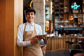 Young happy waiter working in cafe and looking at camera.