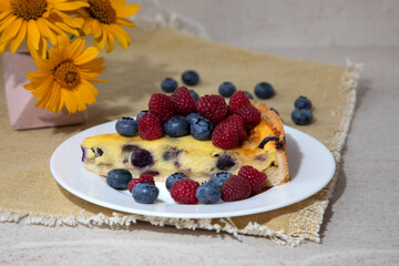 A piece of blueberry cheesecake on a white plate with fresh raspberries and blueberries and yellow flowers on a stone table. Front view