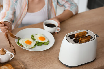 Beautiful young woman drinking coffee and eating tasty toasts with fried eggs in kitchen