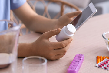 Young man with pill bottle and tablet computer at table in kitchen, closeup