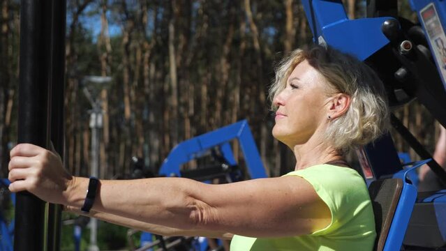 Middle-aged lady does strength training for upper body against blurry trees. Mature blonde woman enjoys workout on sports ground trainer closeup