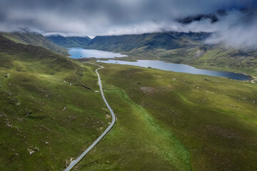 Beautiful scene with small road going into stunning mountains between lush green fields. Connemara, Ireland. Blue cloudy sky. Irish landscape. Transport and travel industry. Aerial view.