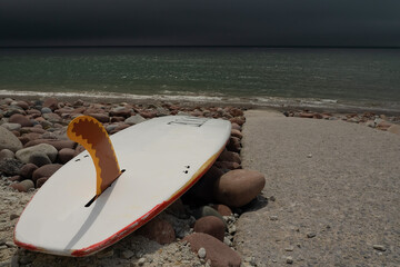 White surfboard on a rough stone shore and path to the ocean with dark storm or hurricane clouds. Stone on the beach has brown color. West of Ireland.