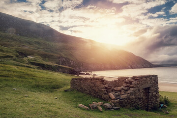 Small old stone building by Keem bay and beach at sunrise. Stunning Irish nature landscape scene. Popular travel area with amazing view. Ireland. Sun flare and cloudy sky. County Mayo, Ireland.