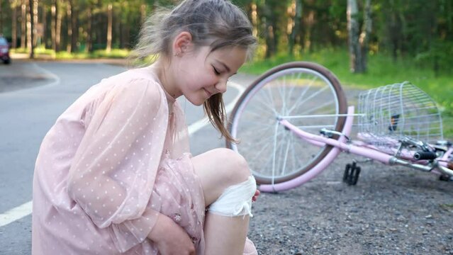 Teen girl grimaces because of pain in leg. Girl wearing pink dress falls from pink bicycle trying to learn to ride bike. Schoolgirl has injury on knee closeup