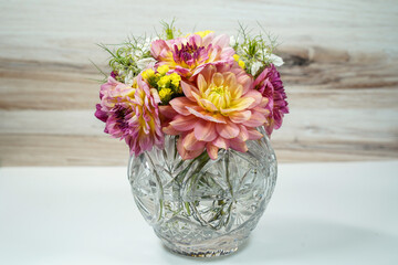 Round glass bowl filled with a bouquet of dahlia and statice flowers. Pink, purple, yellow, and white flowers.  White table with wood background.