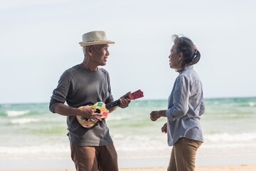 Happy senior couple relaxing outdoors singing and playing acoustic guitar at beach near sea sunny day, Mature man playing ukulele for his wife at sea, plan life insurance at retirement couple concept