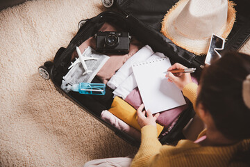 Making check list of things to pack for travel. Woman writing paper take note and packing suitcase to vacation writing paper list sitting on room, prepare clothes into luggage, Travel vacation travel - obrazy, fototapety, plakaty