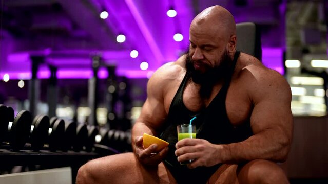 muscular bodybuilder with fruit smoothie and grapefruit in hands is sitting in gym