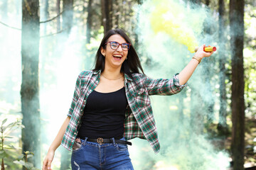 Young woman in a photoshoot raising her hand holding a smoke bomb yellow in the forest smiling at...