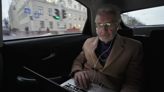Portrait of gray-haired designer with glasses looking at a laptop screen, a focused architect works on the project sitting in the moving car, slow motion.