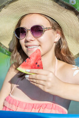 Happy teen girl wearing broad-brim and sunglasses eats watermelon in the pool at sunny day