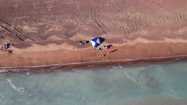 A beach with blue water color and sandy waves. Top view from a drone of vacationing people, tents and cars. Brown sand of Lake Balkhash. Blue sky. Bushes and several trees grow. Family vacation by sea