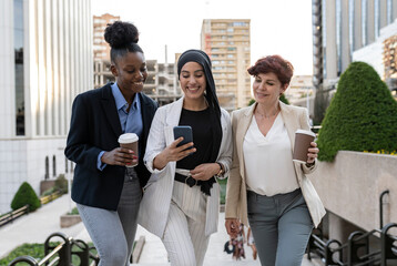 three multiracial female businesswomen of different ages walking outdoors with tablet and coffee,...