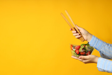 Woman holds salad with strawberry and wooden tongs on yellow background