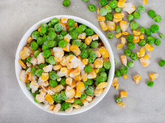 Mix of frozen garden peas and corn in round white bowl on light grey surface