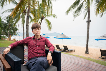 Portrait of handsome asian man red shirt siting on chair and posting over beach background.
