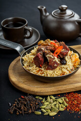 rice pilaf with meat in a frying pan on a board on a black background with a teapot, vertical photo
