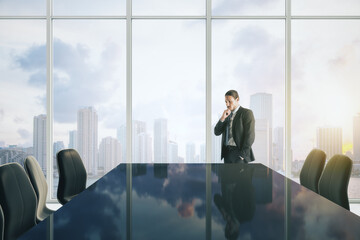 Attractive young european businessman thinking in modern meeting room interior with reflections on table, chairs and panoramic window with sky clouds view and daylight. CEO and conference concept.