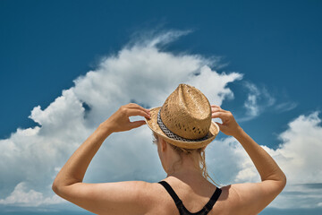 Adult European woman is on the seashore and holds a straw hat on her head with her hands, a silhouette of a beautiful blonde against the background of cumulus clouds over the sea