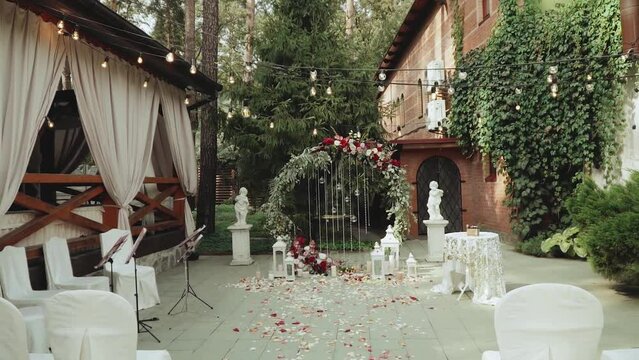 Wedding arch with red flowers and white chairs, lamp garland over the ceremony near restaurant slow motion shot. Boho-chic wedding style.