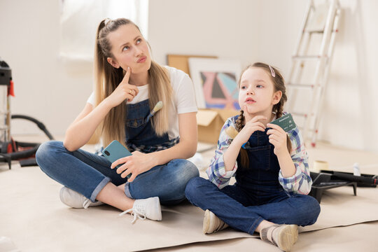 Blonde girl aged 27, together with cute 8-year-old daughter, are sitting on floor. The girls look up thoughtfully. In background there is ladder vacuum cleaner paint rollers.