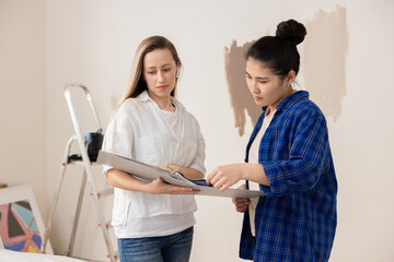 Two young women, one dressed in a white t-shirt and a white shirt, the other in a beige t-shirt and a kilt shirt, stand in the middle of a room undergoing renovations. Behind them, there is a ladder