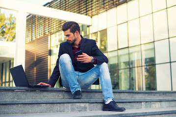 Young businessman using phone and laptop while sitting on the stairs in front of corporate office building